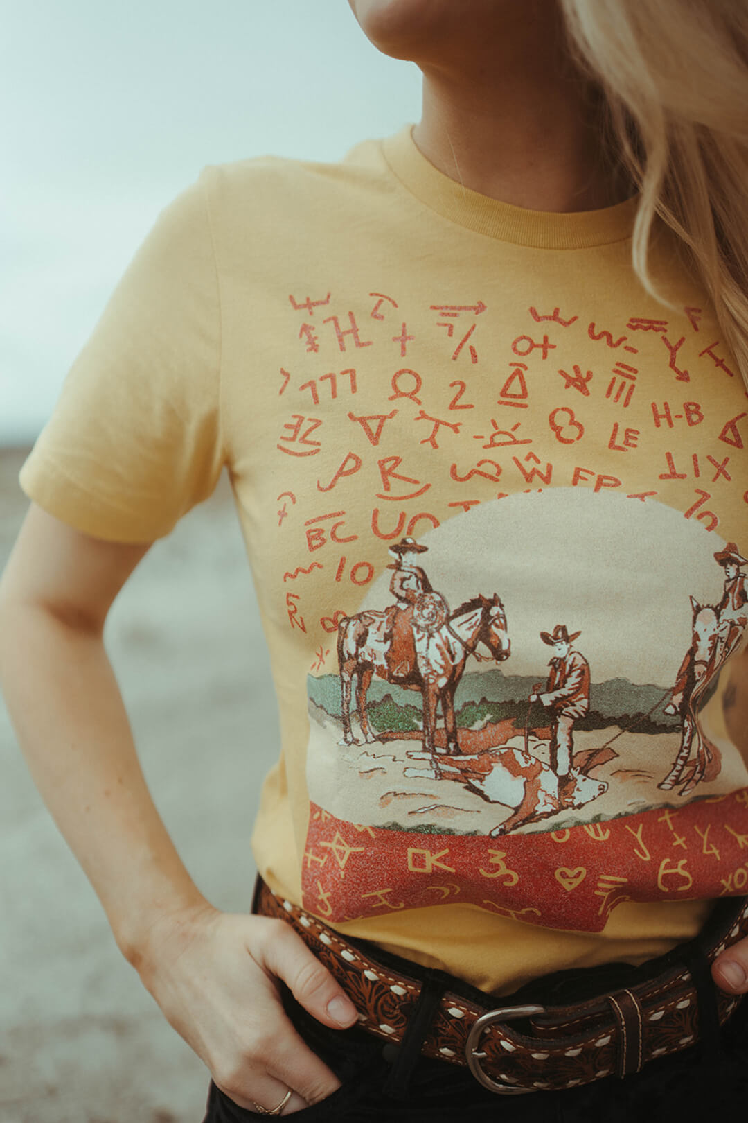 Close up detail picture of yellow color graphic tee from XOXO Art Compnay featuring branding images and cowboys branding a cow. 
